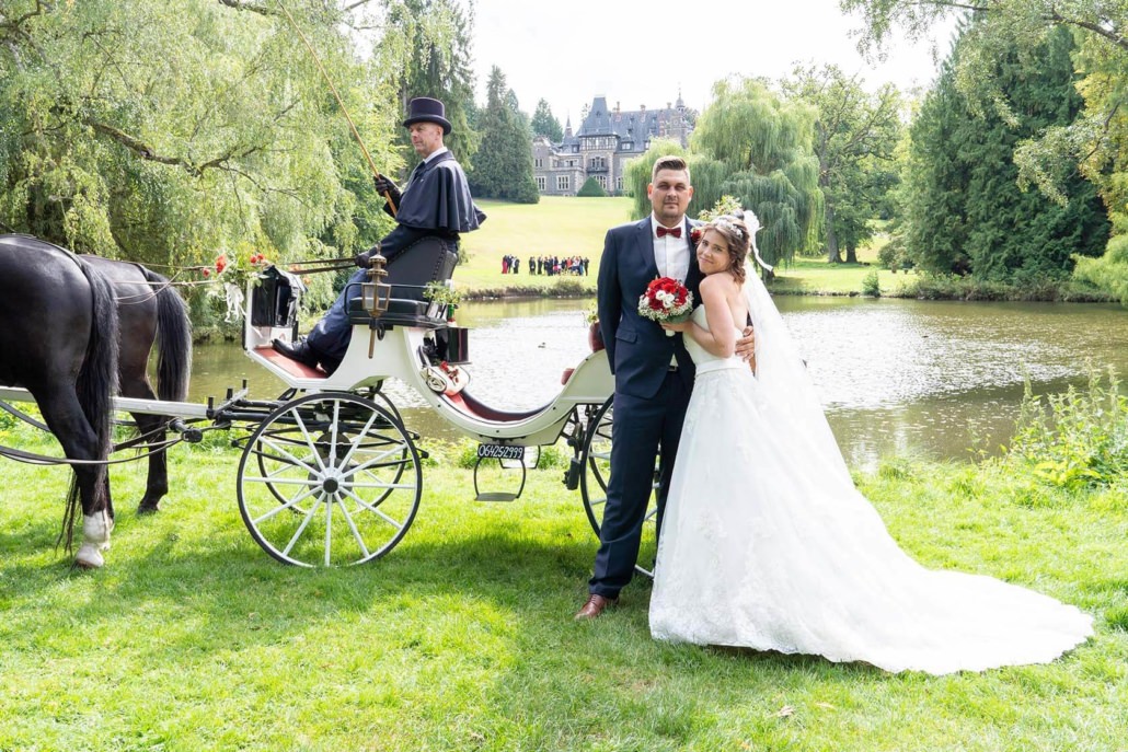 Hochzeitspaar vor der Hochzeitskutsche mit der Hochzeitsgesellschaft und Schloss im Hintergrund